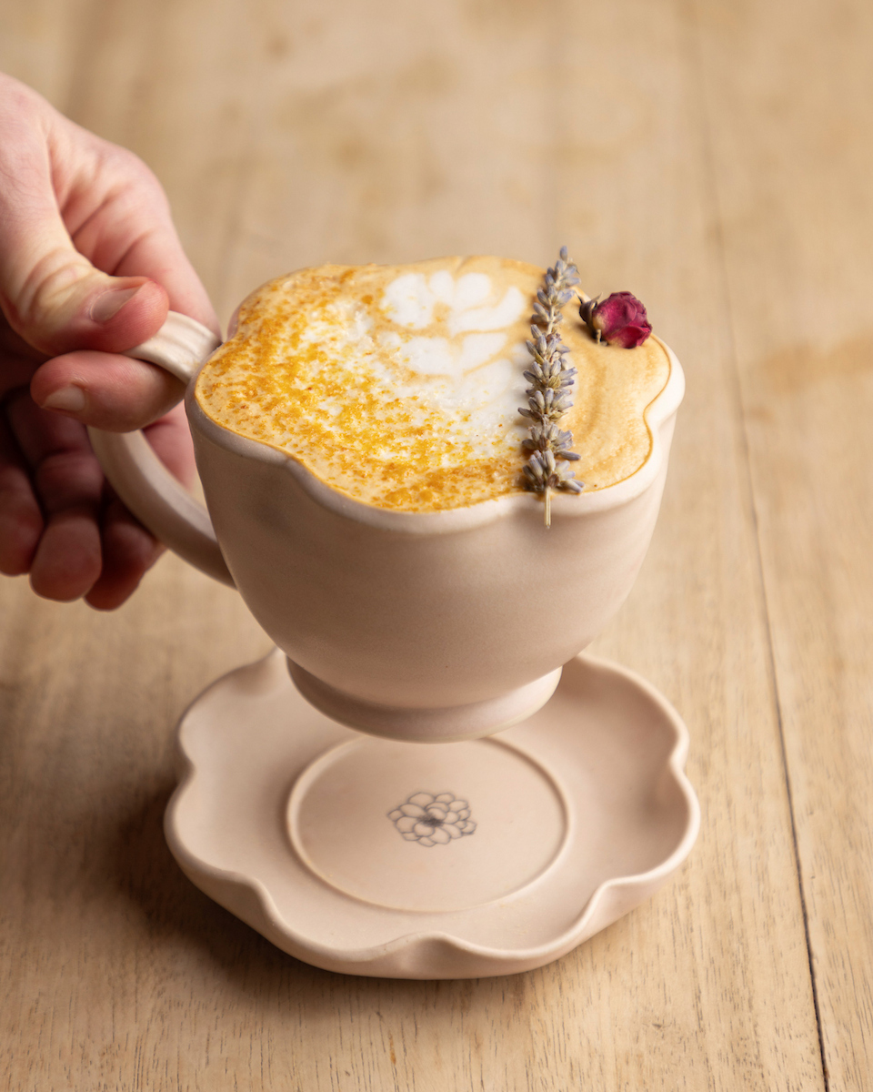 A hand reaching for an artfully crafted latte in a ceramic cup, garnished with lavender and a rosebud